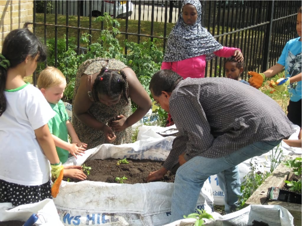 A group of people, including children and adults, engaging in a community gardening activity. A woman with braided hair and a man in a checkered shirt are carefully tending to a raised garden bed filled with soil and young plants. A young girl wearing a patterned hijab and a pink jacket observes the activity in the background. The setting is outdoors, enclosed by a black metal fence with greenery growing around it.