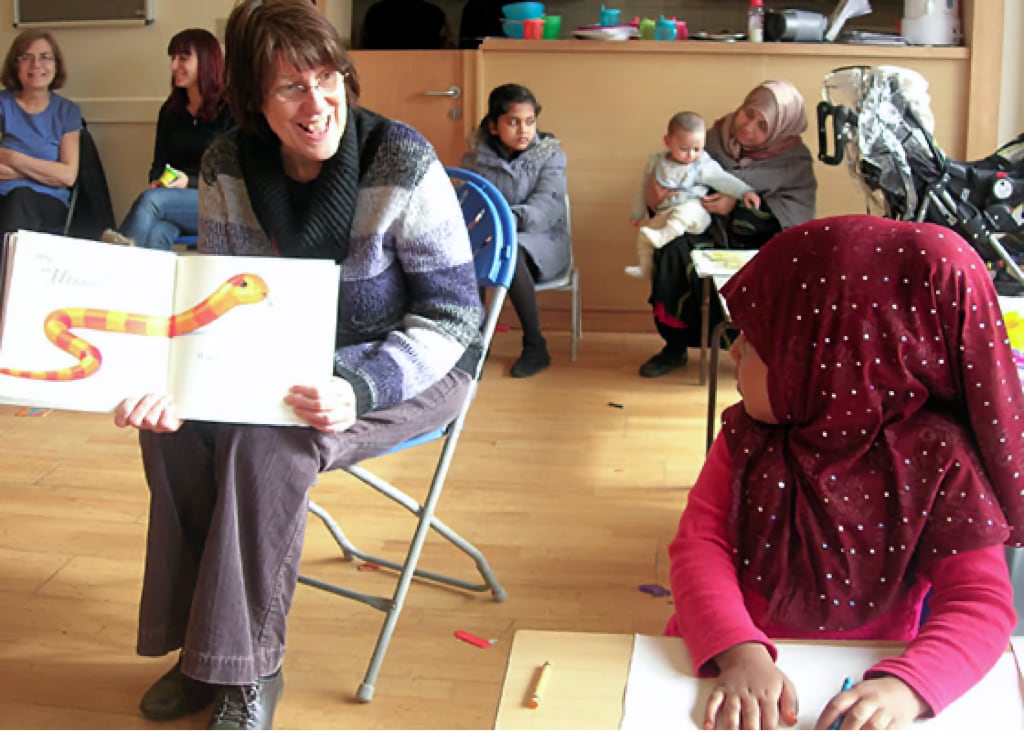 A woman enthusiastically reading a children's book to a young girl in a community setting, with other parents and children engaging in activities in the background.