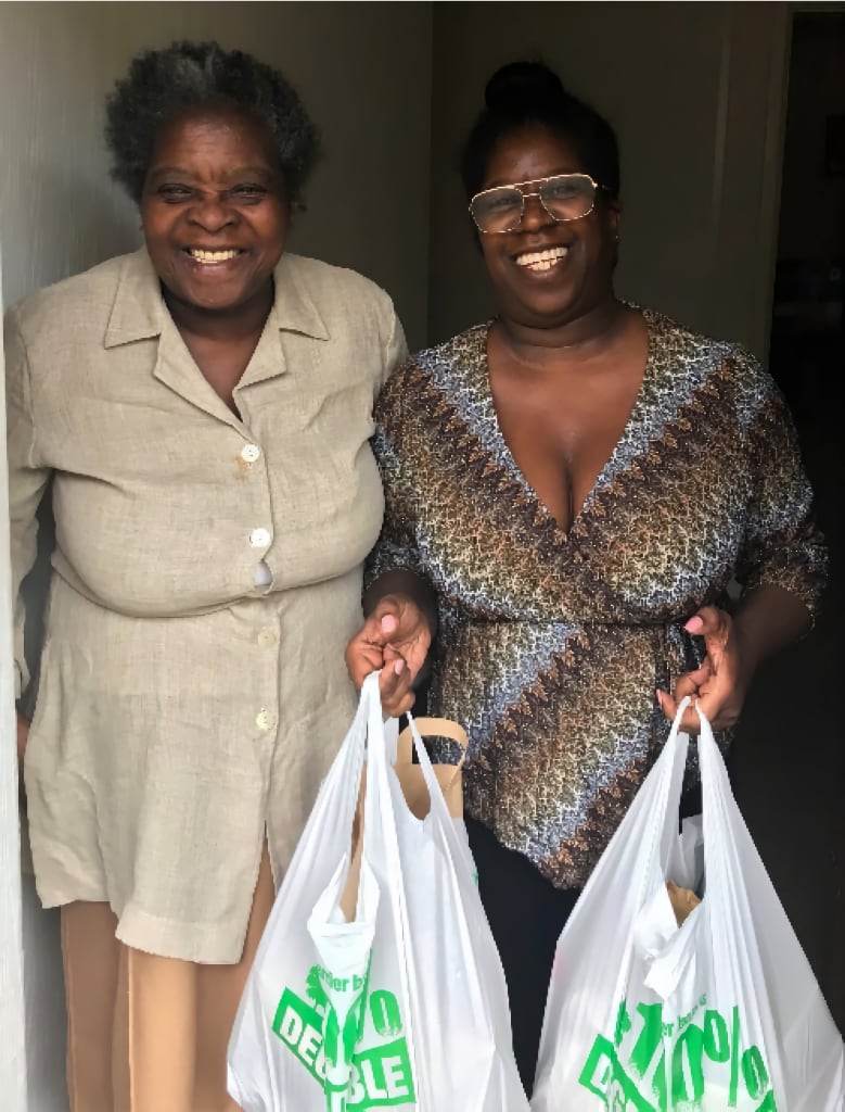 Two smiling women stand in a doorway holding bags of groceries, symbolising community support and volunteer efforts. The image reflects the impact of volunteers in delivering essential items and assisting those in need.