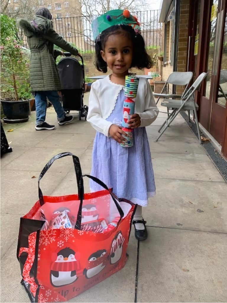 A young girl wearing a handmade festive crown stands outside holding Christmas crackers, next to a holiday-themed gift bag filled with treats, smiling joyfully.