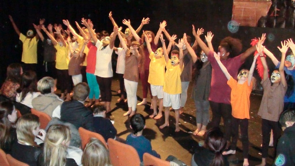 A group of children performing on stage, dressed in brown, white, and yellow outfits with painted faces, raising their arms in unison. The audience watches attentively from orange theatre seats. The performance takes place in a dimly lit theatre, highlighting the expressive gestures of the children. This image represents the impact of community arts and creative expression at Regents Park Time Bank.