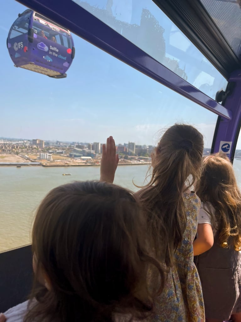 Three young children inside a cable car, looking out over a cityscape with a river below. One child is reaching out towards another passing cable car, which has a "Selfie in the cloud" advertisement on it. The bright blue sky and modern buildings in the distance create a sense of adventure and exploration.