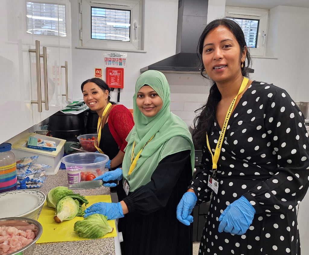 Three women wearing yellow lanyards and gloves prepare food in a kitchen. One is chopping cabbage, another is holding a bowl of tomatoes, and they are all smiling.