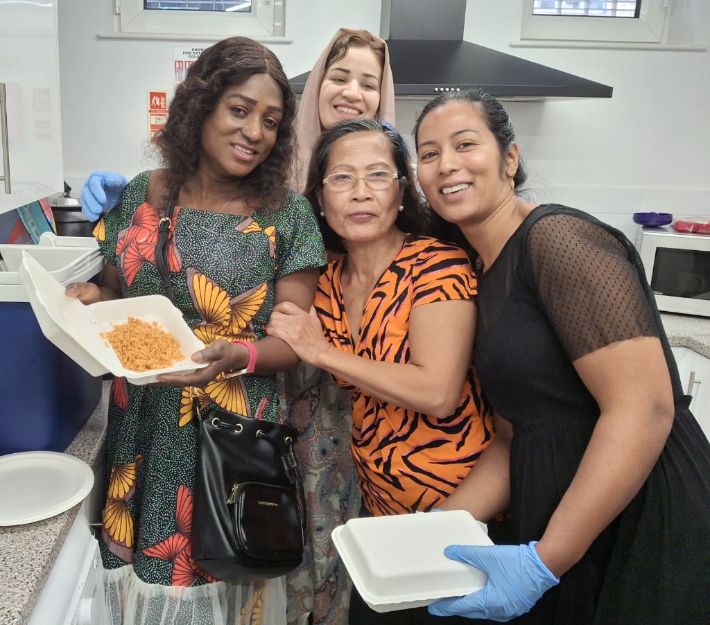 Four smiling women of diverse backgrounds stand together in a community kitchen, embracing each other. Two of them are holding food containers with prepared meals, while one wears gloves, indicating involvement in food preparation. The kitchen setting suggests a communal effort, reflecting the spirit of collaboration and support within the Time Bank initiative.