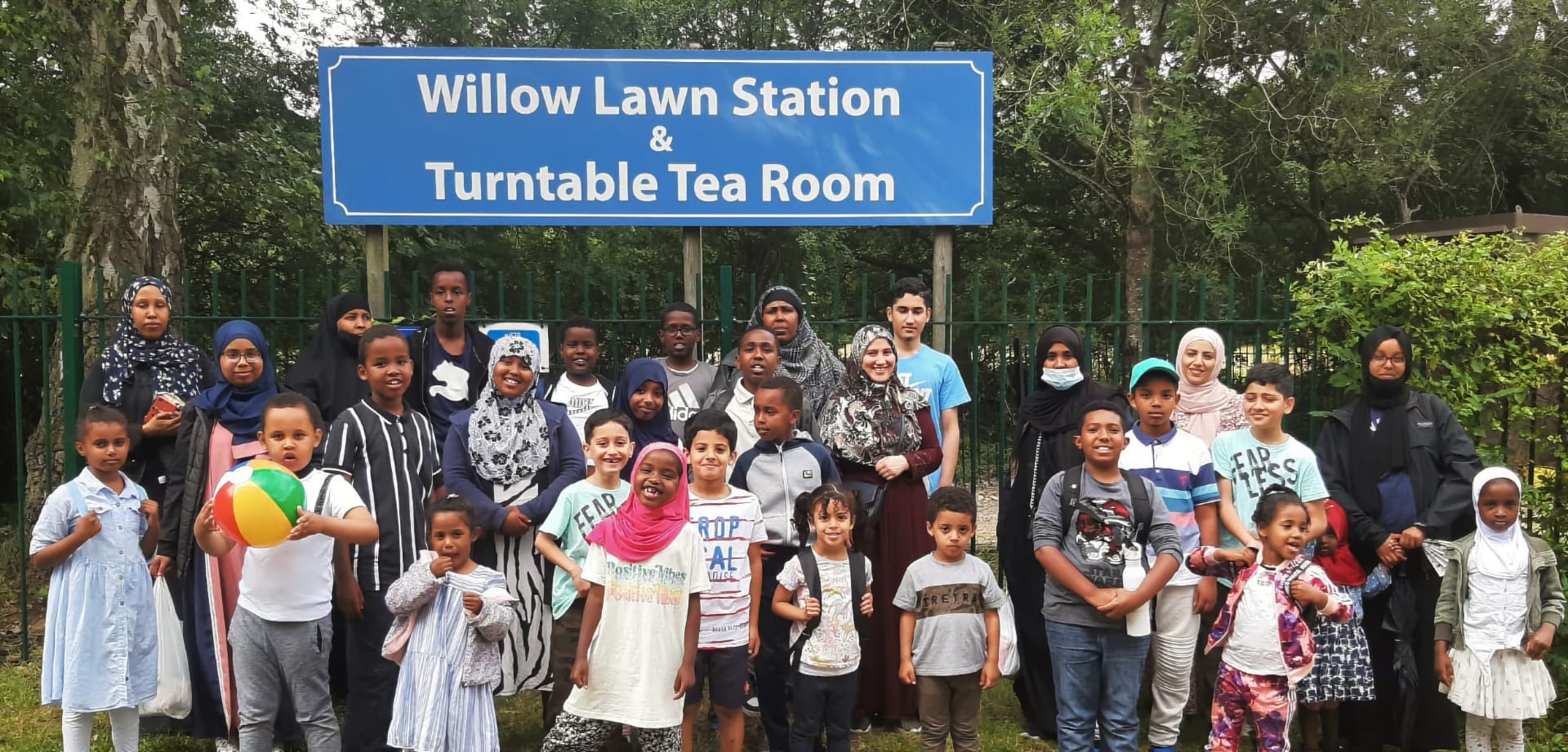 A large group of children and adults gather in front of a blue sign reading "Willow Lawn Station & Turntable Tea Room." The diverse group, featuring families and community members of various backgrounds, stands together outdoors, some holding toys and bags. The image captures a moment of joy and unity during a community trip or excursion.