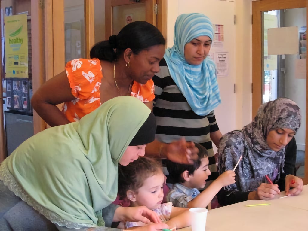 A diverse group of women and children gathered around a table, engaging in a creative activity. Two women, one wearing a green hijab and another in a blue and white hijab, assist young children as they draw and colour. A woman in an orange floral dress leans in with a warm expression. The scene reflects a welcoming and supportive community, embodying the spirit of Regents Park Time Bank’s work in making the borough a place of sanctuary.