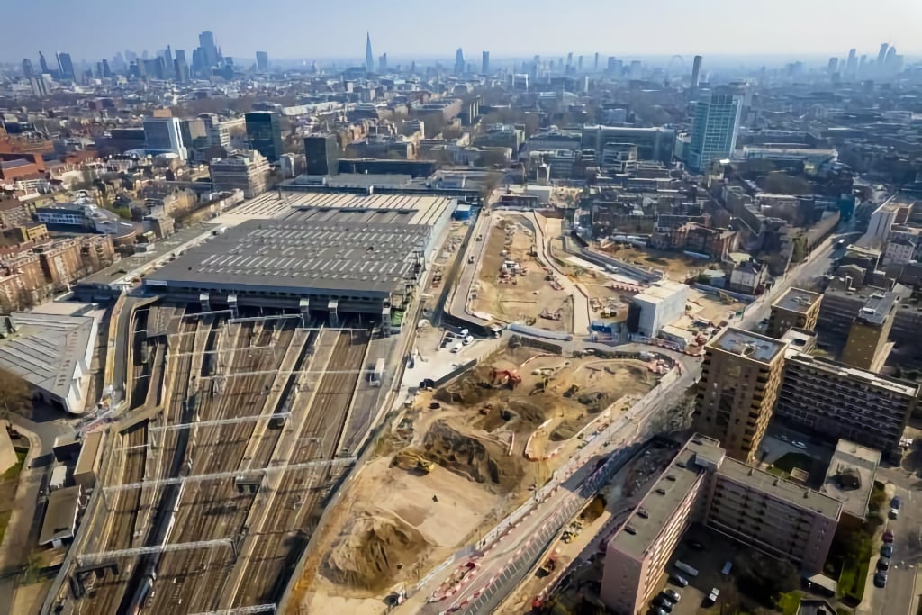 Aerial view of a large construction site next to a railway station in London, with train tracks extending from a massive depot. The construction area features excavated land, heavy machinery, and winding roads under development. The London skyline, including The Shard and other high-rise buildings, is visible in the background. The image captures the scale of urban redevelopment and infrastructure expansion in the city.