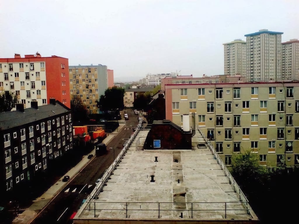 A cityscape view featuring a mix of mid-rise and high-rise residential buildings in an urban setting. The perspective is from a rooftop, showing a street below with parked cars, trees, and a few pedestrians. The buildings display various architectural styles and colours, with older brick structures alongside modern apartment complexes. The sky is overcast, casting a muted light over the scene.