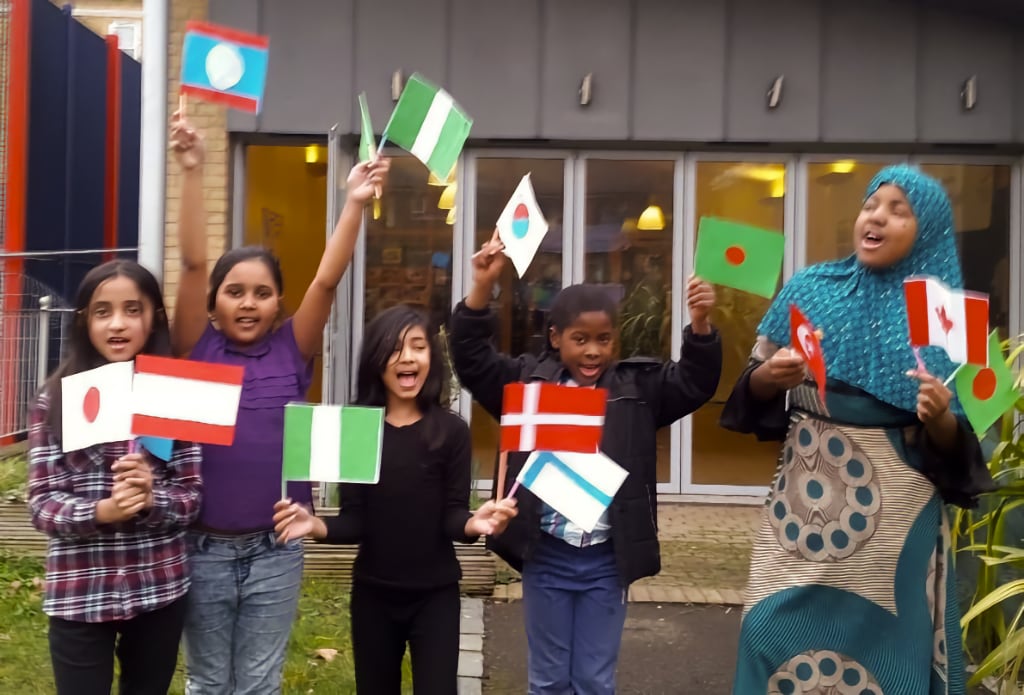 A group of children and an adult joyfully wave small international flags outside a community centre. The children, dressed in casual clothing, hold flags from various countries, including Nigeria, Denmark, and Bangladesh, symbolising cultural diversity and inclusivity. The background features large glass windows and doors, with warm lights visible inside the building.