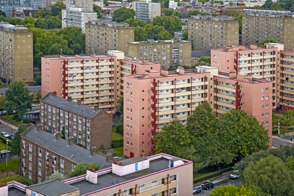 An aerial view of a residential neighbourhood featuring multiple mid-rise apartment buildings with red and beige exteriors. The buildings are surrounded by lush green trees and streets lined with parked cars. The background shows additional housing blocks and a dense urban landscape, representing a vibrant and well-populated community.