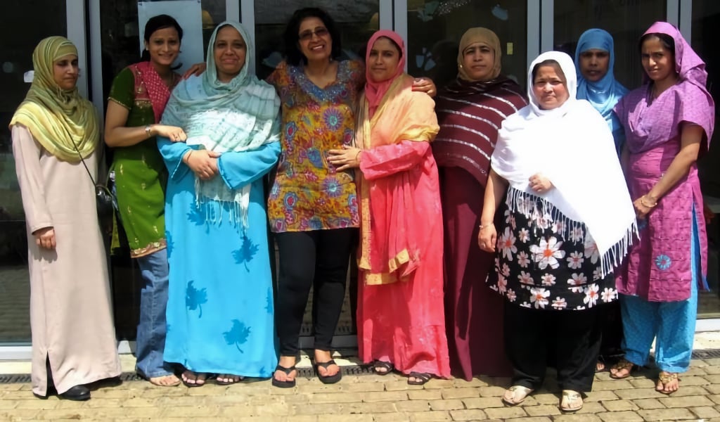 A group of six women standing together outside a building, dressed in a mix of traditional and modern clothing, with warm smiles and arms around each other. They are wearing vibrant and patterned outfits, including headscarves and shawls, reflecting cultural diversity and community spirit. The background features a glass door, suggesting a community centre or gathering place.